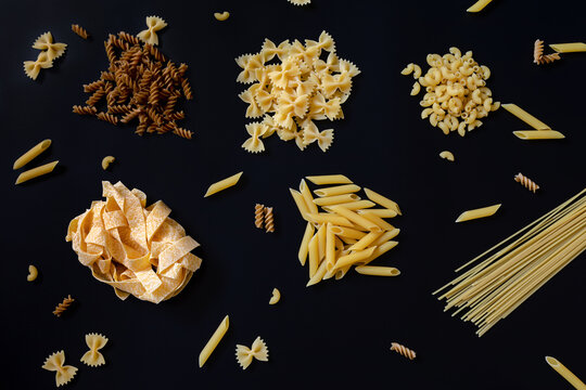 Different types of uncooked dried pasta on the black background. Raw pappardelle, penne, spaghetti, farfalle and fusilli. Italian pasta cooking concept