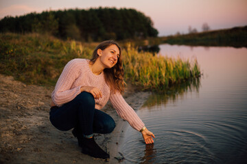 Beautiful girl in a pink sweater sitting by the river. Touch the water with your hand