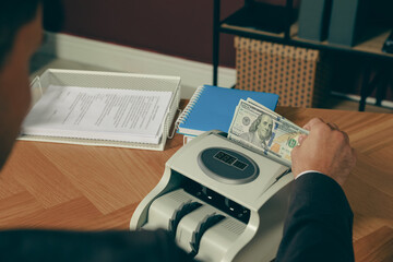 Man putting money into banknote counter at wooden table indoors, closeup