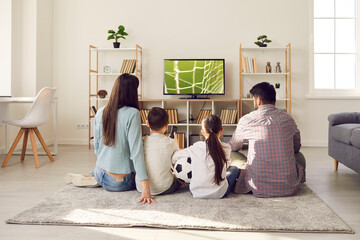 Young family with children watching football match together at home cheering for their favorite team. Mom, dad, daughter and son are sitting on floor in front of big TV with their backs to camera.