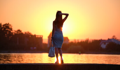 Rear view of lonely woman standing alone on lake shore on warm evening. Solitude and relaxing in nature concept