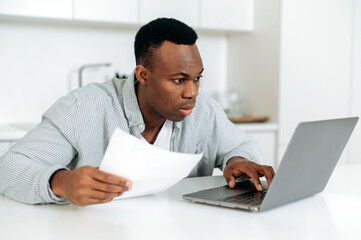Fototapeta na wymiar Concentrated confused young african american man, freelancer, stock trader, sitting at the table at home, in casual wear, in confusion looks into a laptop, analyzes online reporting, profit