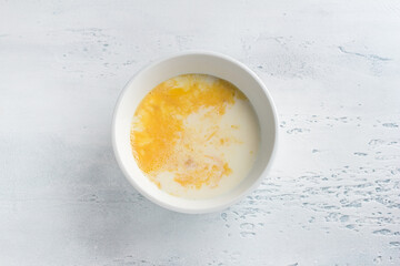 A white bowl with beaten egg and milk on light blue background, top view. Cooking omelet, homemade healthy food