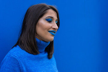 portrait of a smiling young woman with blue eye make-up and lipstick and blue sweater on a blue background.