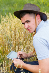 Obraz premium Digital farm. Portrait of farmer seating in gold wheat field and scrolling on tablet. Young man wearing cowboy hat in field examining wheat crop. Oats grain industry. Countryside. Oats plant