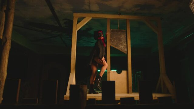 Young sexy woman in mask and black Halloween executioner costume dancing near old vintage wooden guillotine .  Girl with beautiful body with red hair wearing shorts dancing inside dark building . 