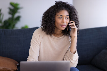 Young black woman at home talking on cellphone using laptop computer sitting on a couch