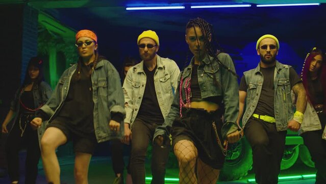 Group of stylish young people dancing synchronized dance  , moving near military tank and police car inside garage building Colorful stylish clothes  Modern professional dances . Green blue led lights