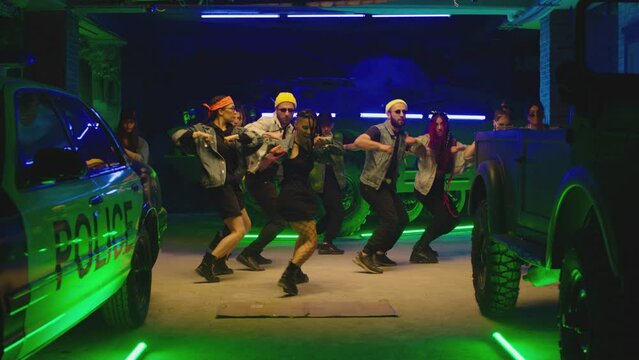Group of stylish young people dancing synchronized dance  , moving near military tank and police car inside garage building Colorful stylish clothes  Modern professional dances . Green blue led lights