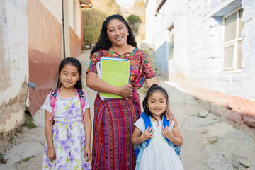 Hispanic mom and daughters ready to go to school - Latin mom accompanying her daughters to school - Hispanic girls with backpack outside their house in rural area - obrazy, fototapety, plakaty