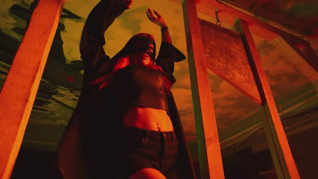 Young sexy woman in mask and black Halloween executioner costume dancing near old vintage wooden guillotine .  Girl with beautiful body with red hair wearing shorts dancing inside dark building . 