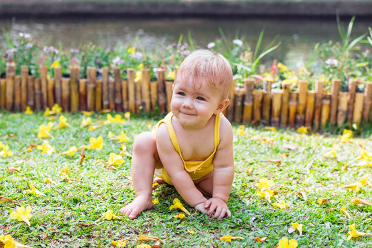 Smiling baby girl with blond hair is playing on grass with yellow flowers on a sunny day in park.