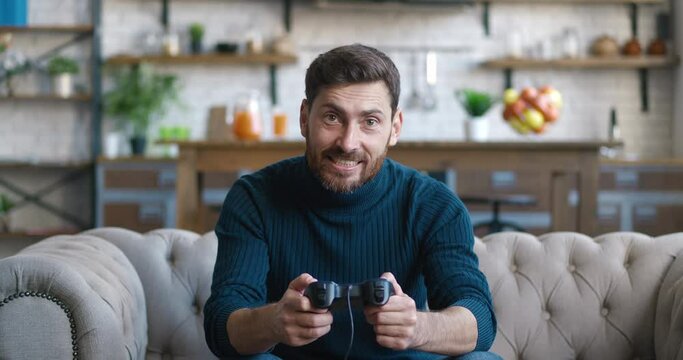 Happy cheerful man with a beard playing console video games using joystick sitting on a sofa at home