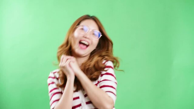 Cheerful young Asian woman with eyeglasses raising hands up, shaking and having fist pump from happiness, smiling woman face, green screen background.