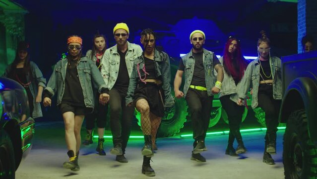 Group of stylish young people dancing synchronized dance  , moving near military tank and police car inside garage building Colorful stylish clothes  Modern professional dances . Green blue led lights