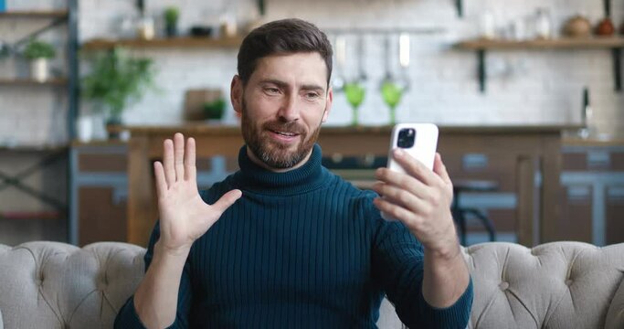 Portrait of man making facetime video calling with smartphone at living room, waving at people on phone screen. Young male blogger making vlog videoblog using mobilephone. Online communication concept