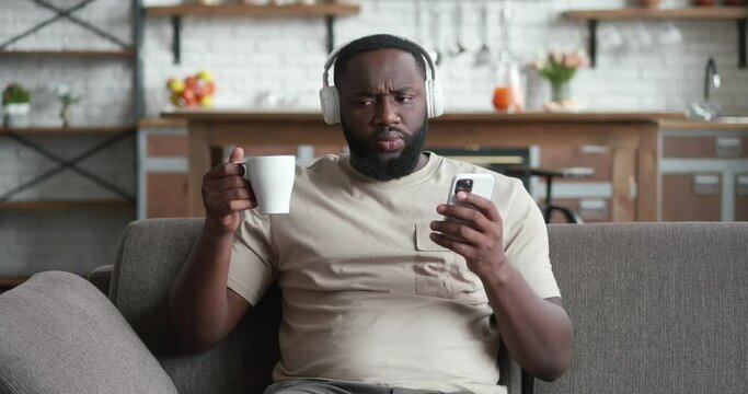Smiling black man in headphones using smartphone sit on couch at home, young african guy drinking coffee holding mobile phone looking at screen reading e-book app listening music online