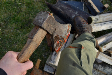 a bloody ax stuck in a log for chopping wood. a man in green pants and leather boots looks at a cut injury on his foot and leg or knee. awkward and combative scene from the rural environment