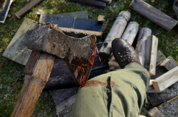 a bloody ax stuck in a log for chopping wood. a man in green pants and leather boots looks at a cut injury on his foot and leg or knee. awkward and combative scene from the rural environment