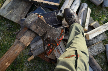 a bloody ax stuck in a log for chopping wood. a man in green pants and leather boots looks at a cut injury on his foot and leg or knee. awkward and combative scene from the rural environment