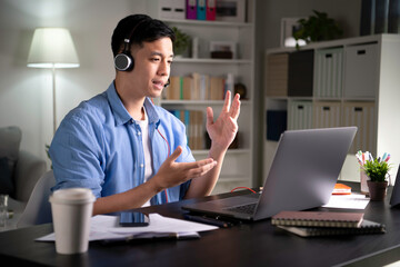 Focused young Asian business person, entrepreneur, speaker, student, office worker wearing headphones, looking at laptop screen and speaking in video conference call, giving lecture, joining online