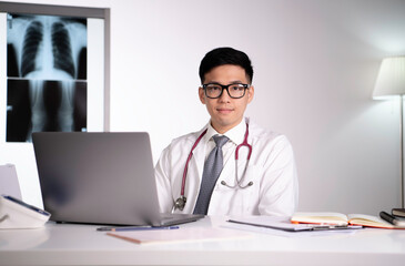Kind and polite young Asian medical doctor orthopaedic surgeon wearing stethoscope sitting with laptop looking straight with x-ray image behind in examination room in hospital. 