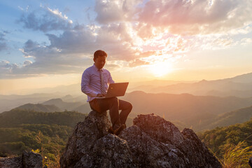 Business man working outdoors with laptop computer on top rock mountain at sunset background. Work and vacation.