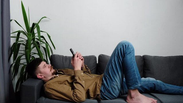 Young man lying on couch hold smartphone spend free time on internet, advanced user of betting, poker and casino gambling websites, online e-commerce buyer. Social media network, modern tech concept