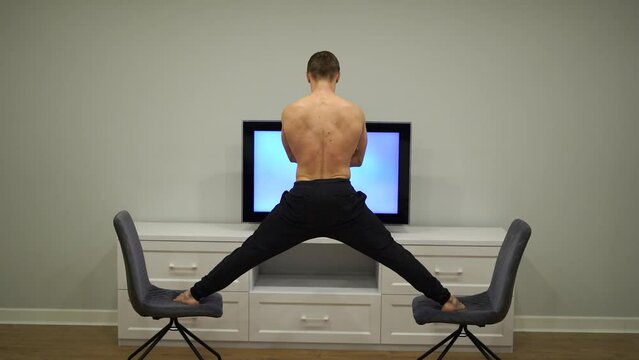 Flexible man keep balance in split between two chairs watching TV. Concept of originality, creativity and outstanding