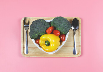 flat lay of vegetables  capsicum, broccoli and tomato in heart shape plate with fork and spoon in wooden tray  on pink background with copy space, healthy lifestyle and weight loss concept.