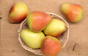 Few red and yellow pears lays in small sandy brown wicker straw basket on kitchen wooden table top view closeup