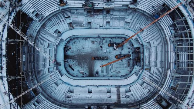 Snowy football field inside stadium with seats for spectators covered with snow and high steel building cranes pull out aerial view