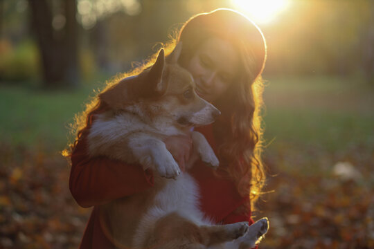 A girl with long red hair, in a red coat and beret gently hugs her corgi dog in an autumn park. Golden autumn, dog walking, friendship, sunset in the park, beautiful autumn picture