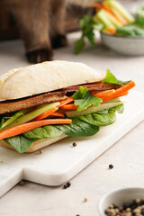 Traditional vietnamese banh mi sandwich with sliced smoked tofu, fresh spinach, shredded carrots and peeled cucumbers, cilantro on marble board on light grey textured background