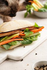 Traditional vietnamese banh mi sandwich with sliced smoked tofu, fresh spinach, shredded carrots and peeled cucumbers, cilantro on marble board on light grey textured background