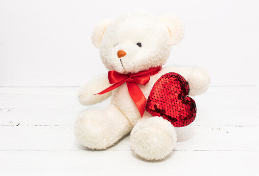 Image of white toy teddy bear sitting on white wooden background with red bow andl heart