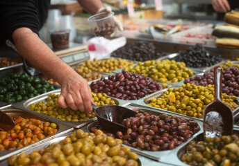 Different colors and taste olives in plastic boxes on stall in foreground. Market. Seller's hand picks up brown olives with scoop. Traditional arabic and jewish food. Mediterranean appetizer. 
