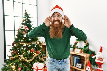 Redhead man with long beard wearing christmas hat by christmas tree trying to hear both hands on ear gesture, curious for gossip. hearing problem, deaf