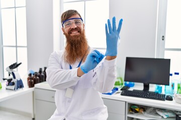 Young redhead man wearing scientist uniform wearing gloves at laboratory