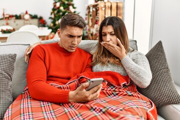 Young hispanic couple sitting on the sofa on christmas covering mouth with hand, shocked and afraid for mistake. surprised expression