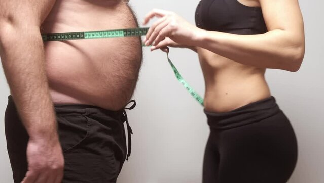 an overweight man measures his waist circumference with a tape measure and shows folds of excess fat on his stomach. Overweight problem.
