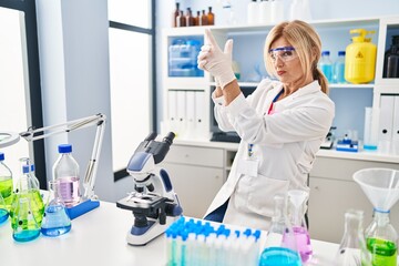 Fototapeta premium Middle age blonde woman working at scientist laboratory holding symbolic gun with hand gesture, playing killing shooting weapons, angry face