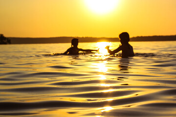 Fototapeta na wymiar Children frolicking playing in water of river, lake, sea at sunset. Preschoolers on summer vacation. Silhouettes of kids have joy on setting yellow sun background. Soft small waves rolling onto shore.
