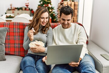 Young hispanic couple watching film eating popcorn sitting on the sofa at home.