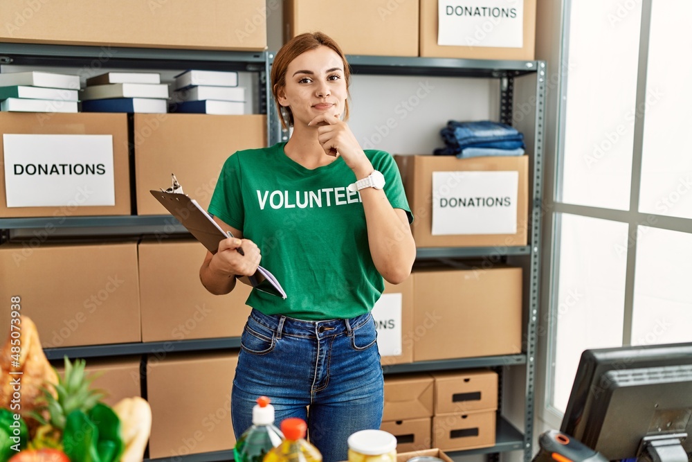 Poster Young brunette woman wearing volunteer t shirt at donations stand looking confident at the camera smiling with crossed arms and hand raised on chin. thinking positive.