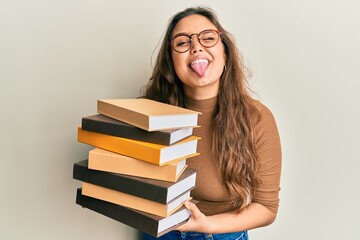Young hispanic girl holding a pile of books sticking tongue out happy with funny expression.