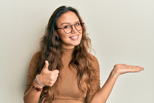 Young hispanic girl wearing casual clothes and glasses showing palm hand and doing ok gesture with thumbs up, smiling happy and cheerful