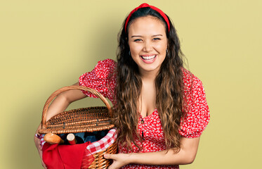 Young hispanic girl holding picnic wicker basket with bread smiling and laughing hard out loud because funny crazy joke.