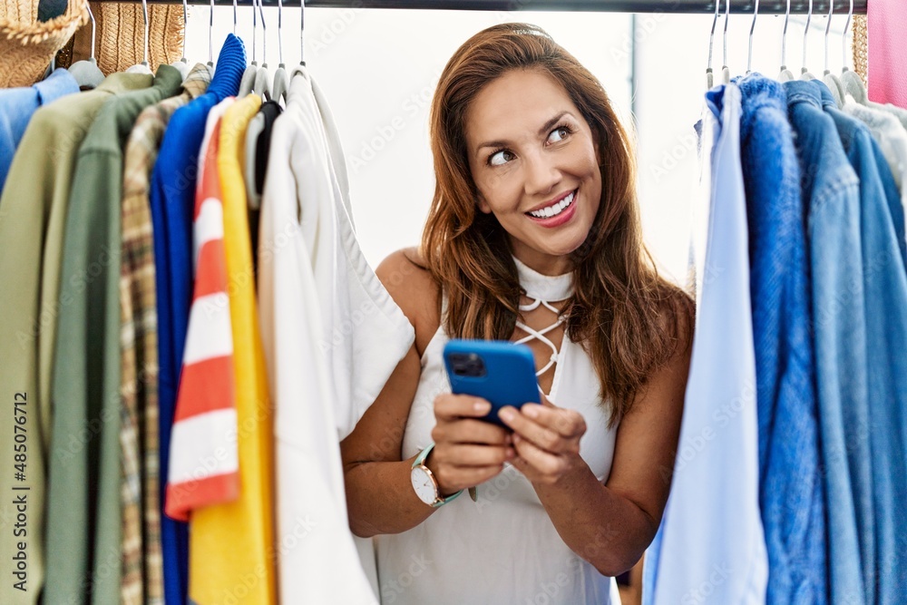 Sticker young latin woman customer using smartphone appearing through clothes rack at clothing store