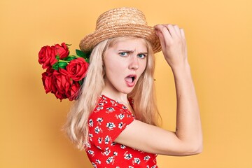 Beautiful caucasian woman with blond hair holding bouquet of red roses in shock face, looking skeptical and sarcastic, surprised with open mouth